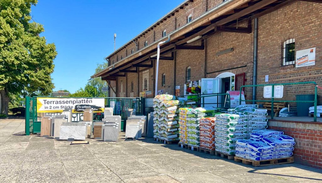 Ein Baumarkt mit gestapelten Baumaterialien, an einer Backsteinfassade auf einem sonnigen Tag.