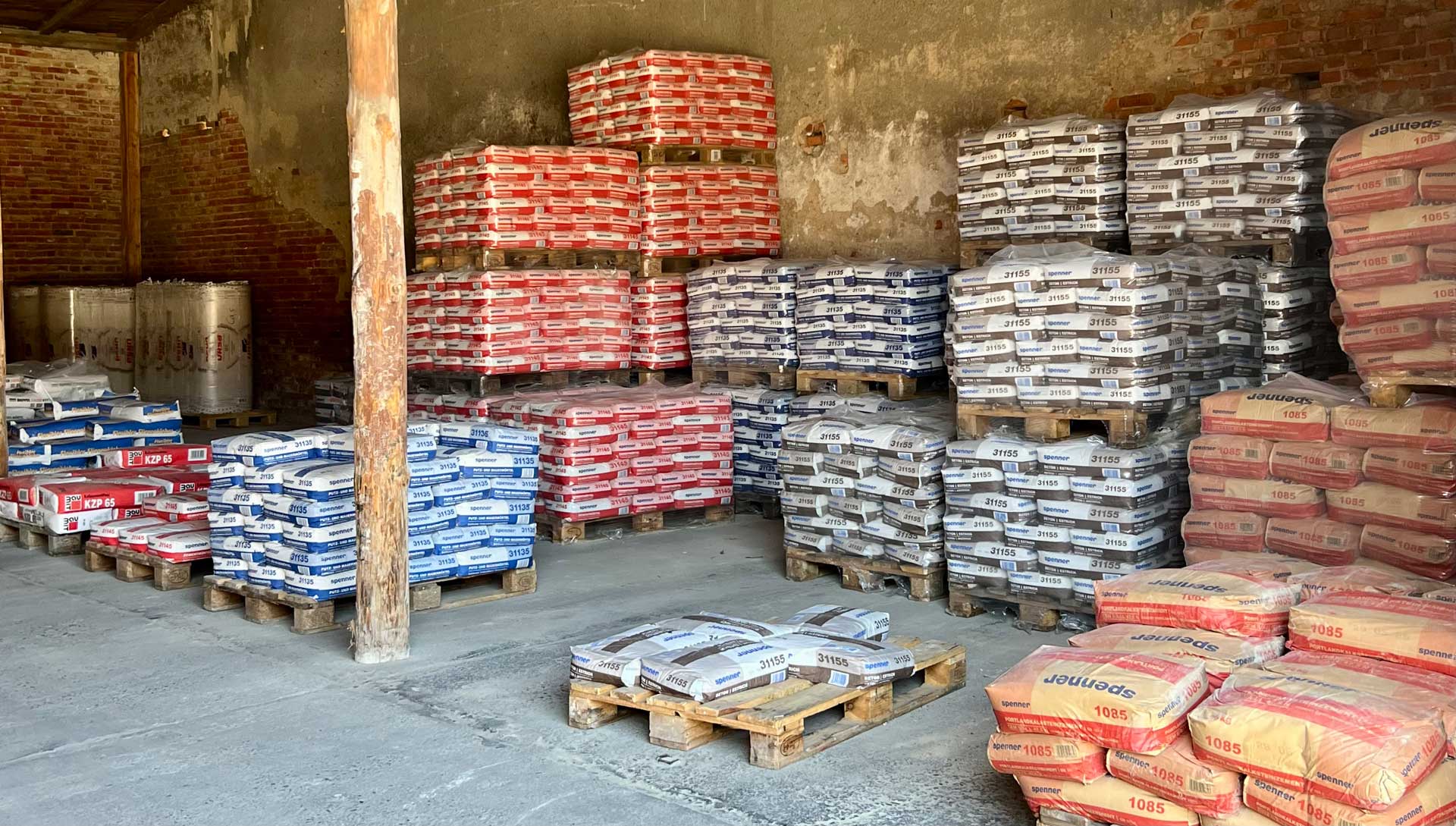 Mehrere gestapelte Paletten mit verpacktem Baumaterial in verschiedenen Farben in einem Lager.