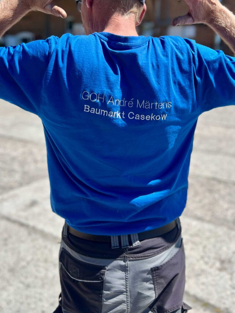 Rückansicht eines Mannes in blauem T-Shirt mit Aufschrift, hebt Arme, sommerliche Umgebung.